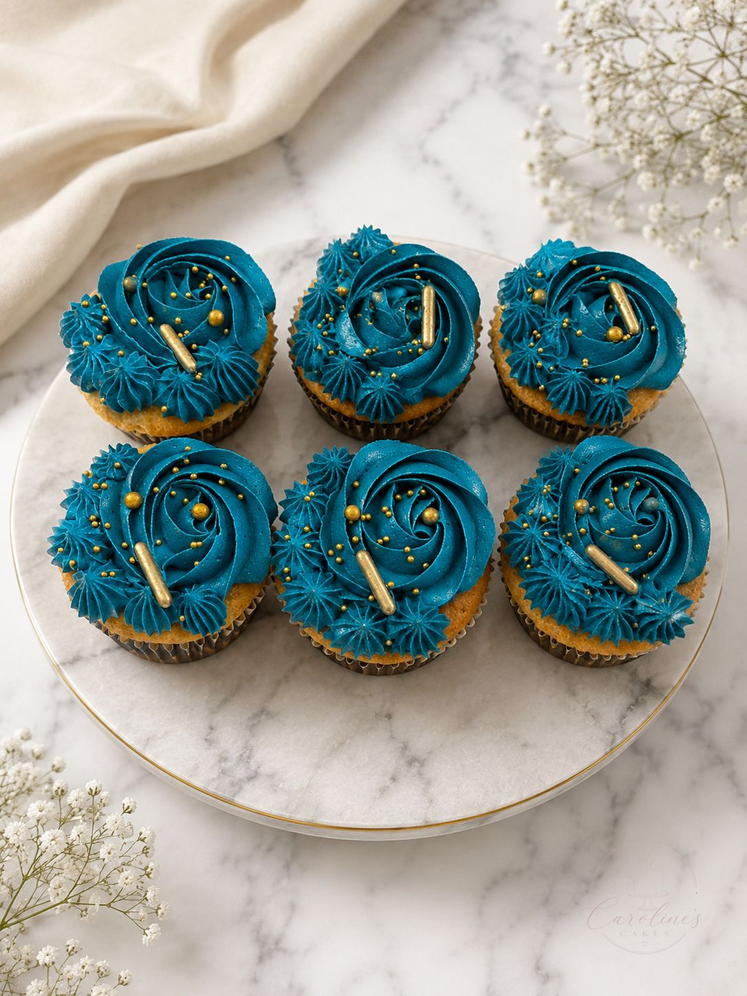 Teal blue buttercream cupcakes with gold pearls and sprinkles on a marble cake stand